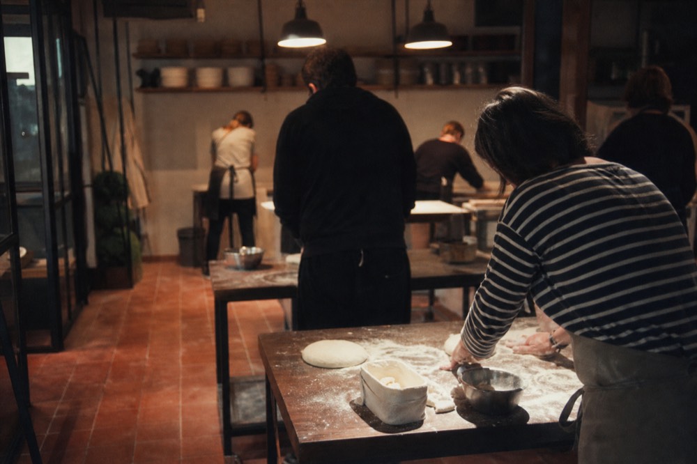 Adults at flour-covered stations in a warm terracotta-floored kitchen, shaping dough under pendant lights