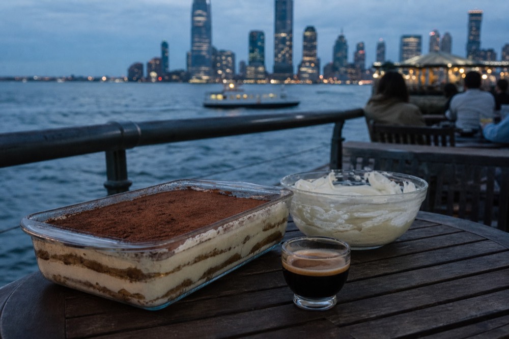 Glass dish of tiramisu dusted with cocoa beside a bowl of mascarpone and an espresso cup