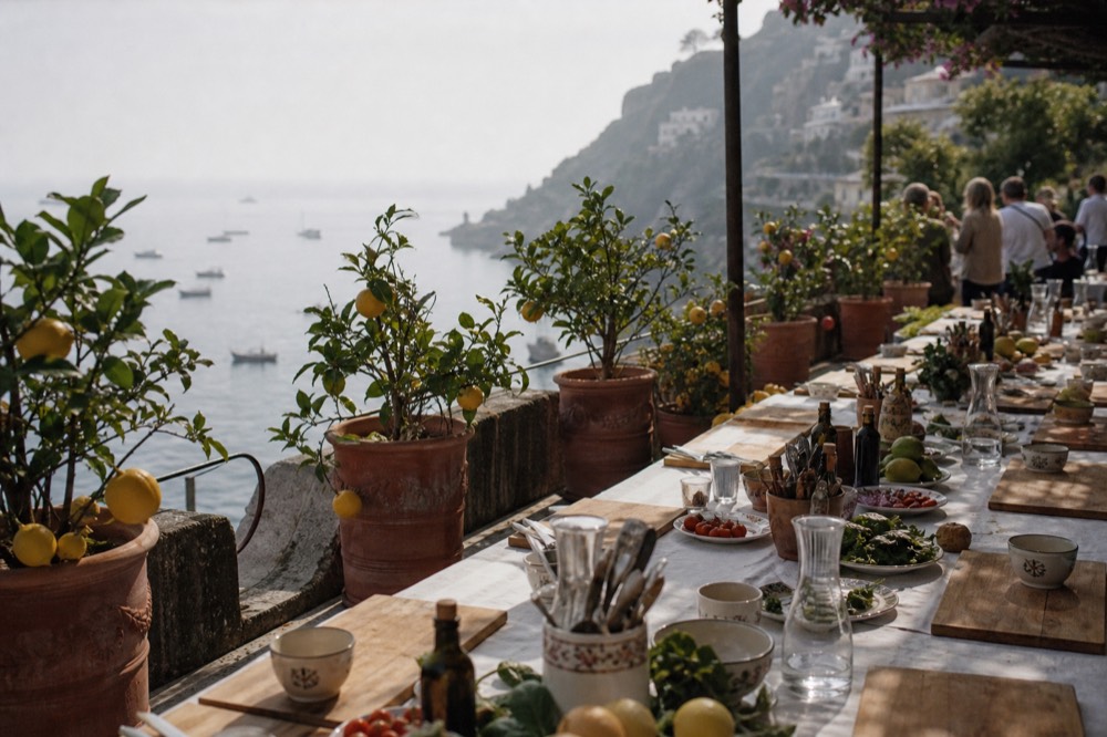 A long terrace table set for a cooking class meal above a steep coastline, lemon trees in terracotta pots and the sea hazy in the distance