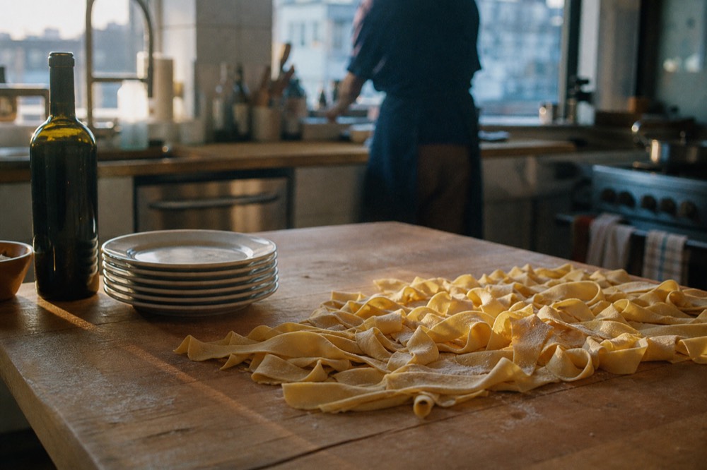 Wooden board of finished handmade pappardelle beside a stack of clean plates and a bottle of red wine in a sunlit kitchen