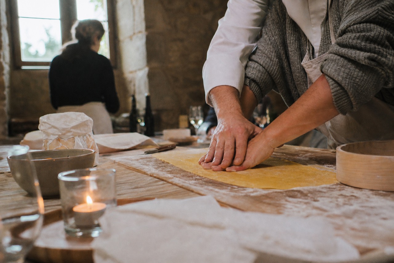 Chef's hands guiding a guest's hands over a sheet of egg dough on a floured wooden board in a warm stone kitchen