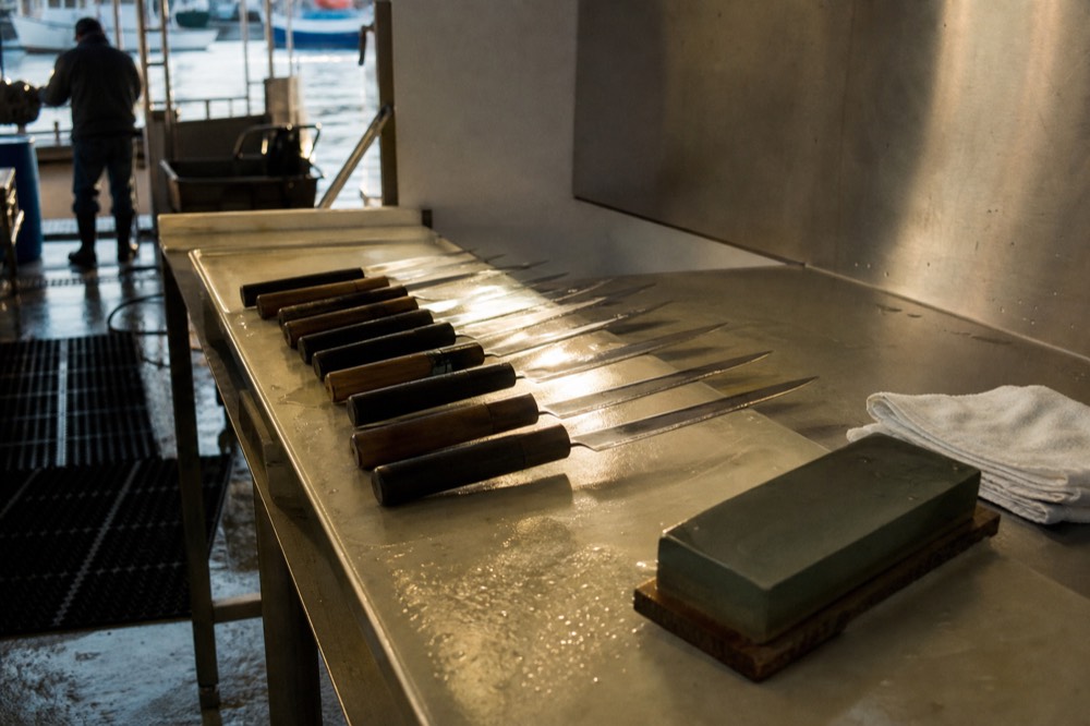 A row of chef knives laid out on a steel bench beside a whetstone and a folded white cloth under bright kitchen light