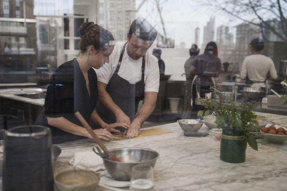 A chef's hands guiding a guest's hands over a rolled pasta sheet on a marble bench, a small group blurred in the background