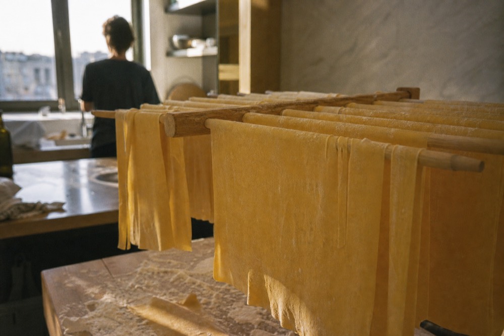 Sheets of fresh egg pasta draped over a wooden drying rack in a bright kitchen, flour scattered across the bench below