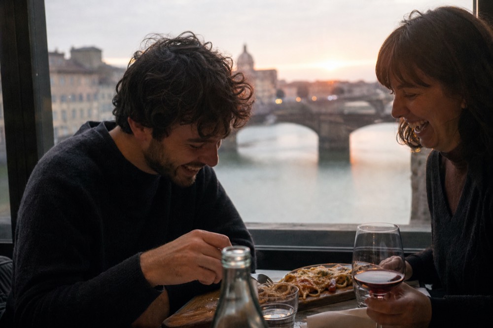 Two people laughing over a shared pasta board by a window with the Arno and an old stone bridge blurred behind them at dusk