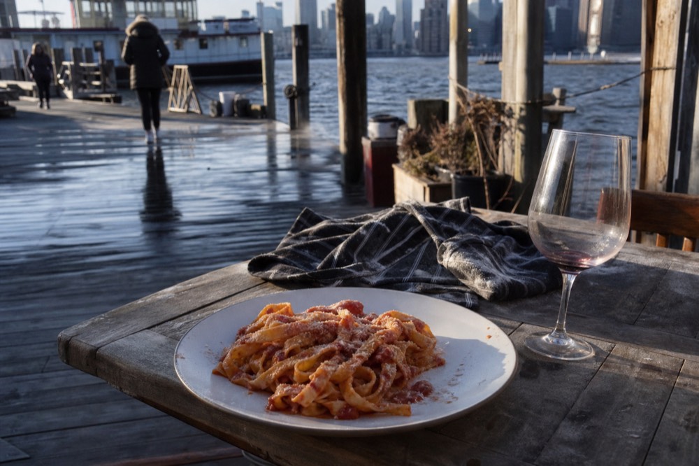 Plate of handmade tagliatelle in tomato sauce set down on a rustic table beside a nearly empty wine glass and a flour-smudged apron