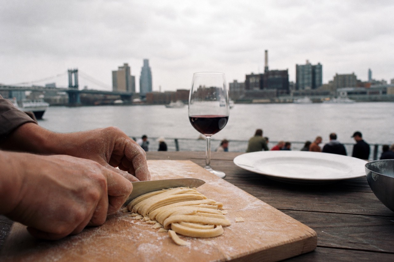 Pair of floury hands cutting a rolled sheet of pasta into ribbons on a wooden board, wine glass and empty plate waiting behind