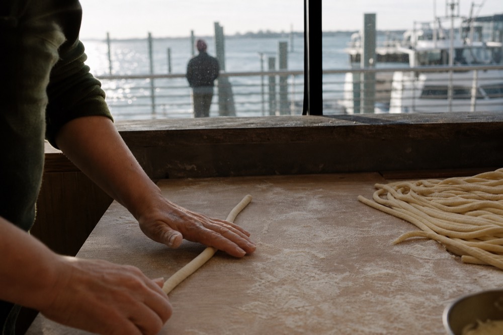 A pair of hands rolling a thick rope of pici between the palms on a floured board, more ropes drying in a coil beside them