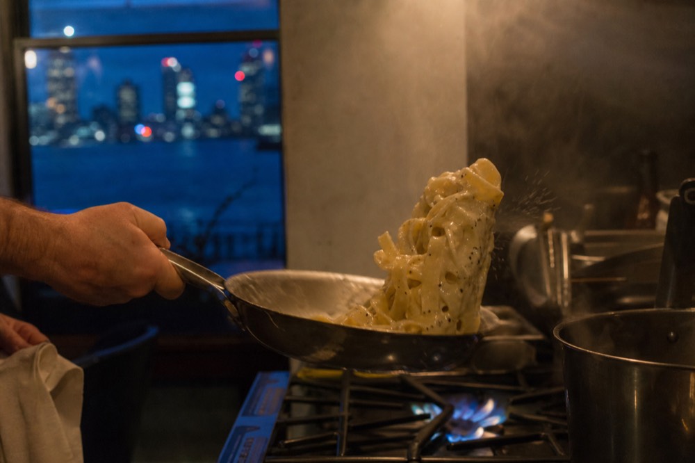 A steel pan of pasta being tossed with a pale cheese emulsion, black pepper visible, steam catching low kitchen light