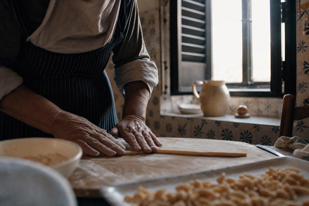 Weathered hands rolling a long rope of pasta dough on a worn wooden board in a small village kitchen with painted tiles and a half-shuttered window