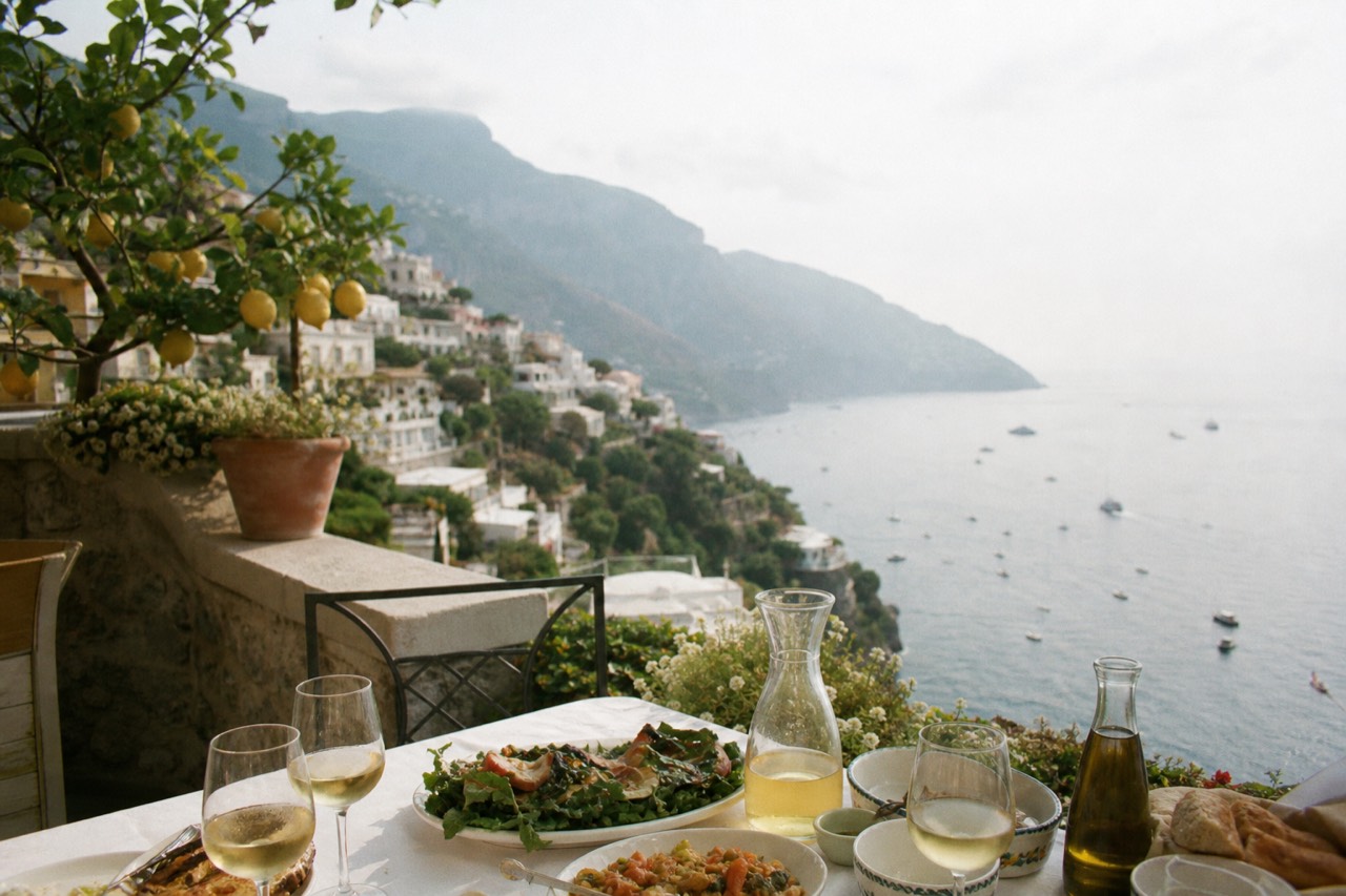 Terrace table above a steep Mediterranean coastline set with platters of food and carafes of wine, lemon trees and hills falling toward the sea