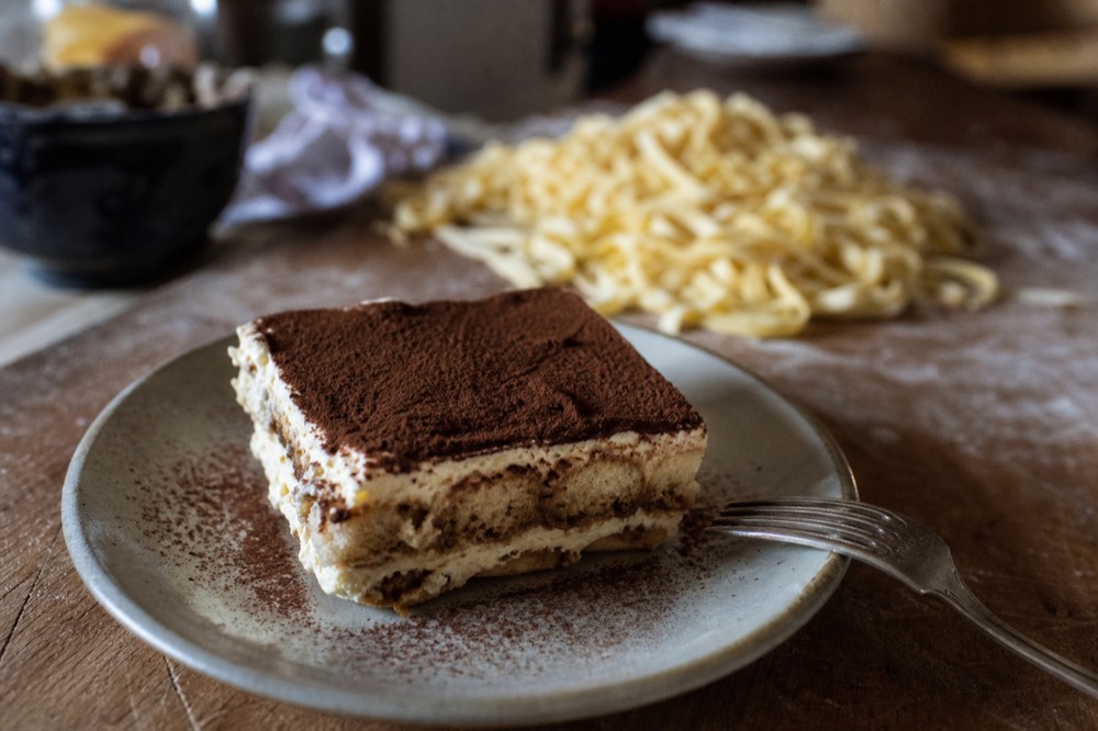 Tiramisu layers being assembled in a glass dish beside fresh pasta