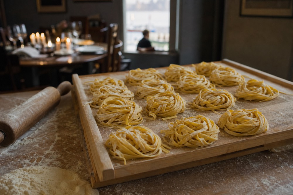 Nests of fresh tagliatelle drying on a wooden rack beside a rolling pin, dusted with semolina, lit by low afternoon light