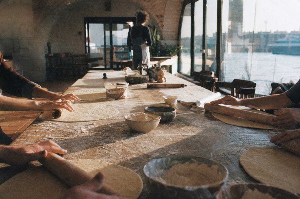 Long communal wooden table with six pairs of hands rolling dough, flour clouds in the air, terracotta floor and whitewashed vaulted ceiling