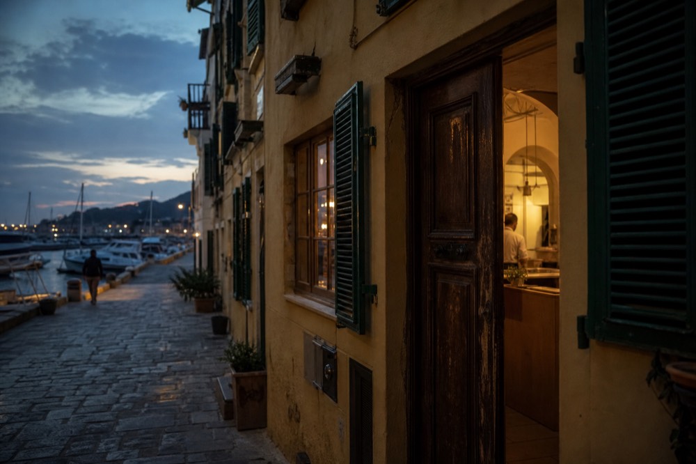 Narrow Florence street of ochre buildings with green shutters, a wooden doorway propped open onto a lit kitchen at dusk