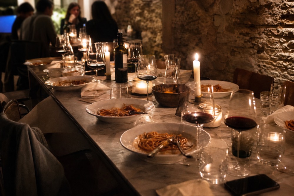 Evening kitchen scene with a long table of finished pasta plates, half-full wine glasses and candlelight against stone walls