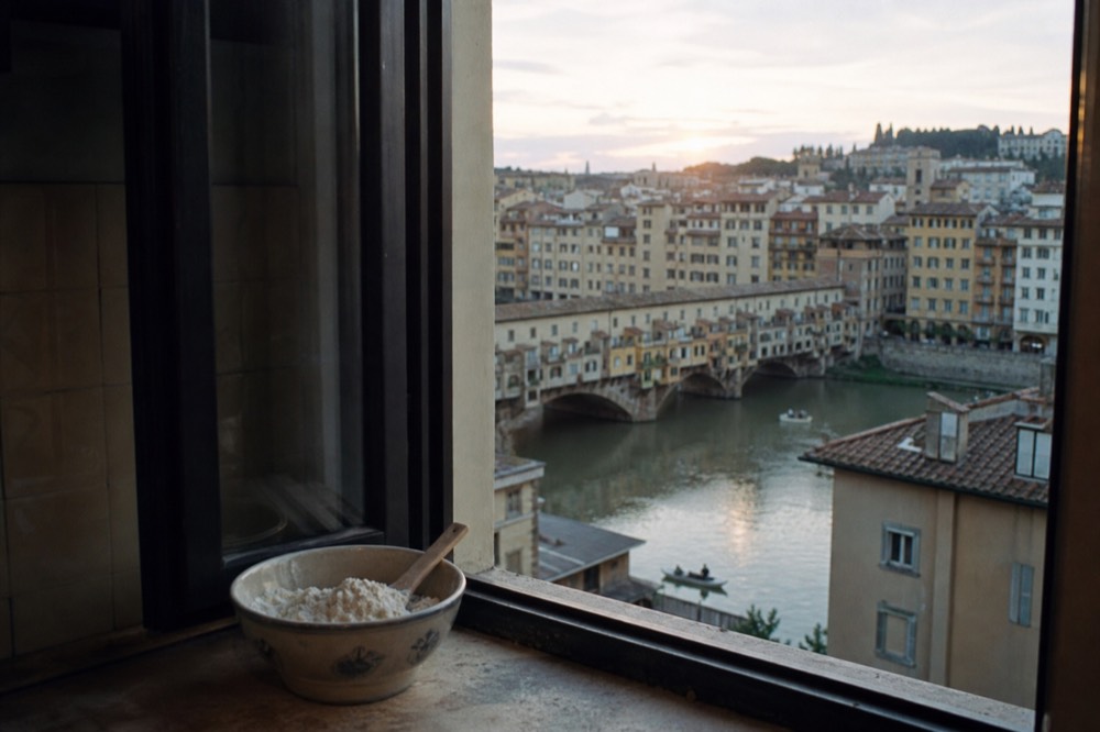 Florence rooftops and the Arno at golden hour seen from a kitchen window, with a bowl of flour on the sill in the foreground