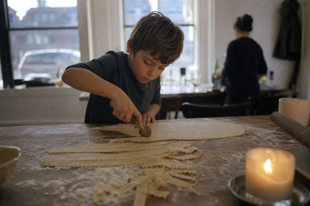 A boy of about eight concentrating hard as he cuts ribbons from a rolled dough sheet with a fluted wheel, flour dusted across the dark wood bench