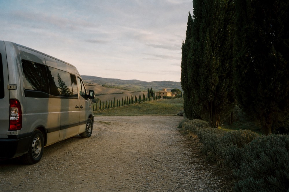 A minibus parked on a gravel track beside cypress trees and rolling Tuscan hills in late afternoon light, farmhouse buildings in the distance