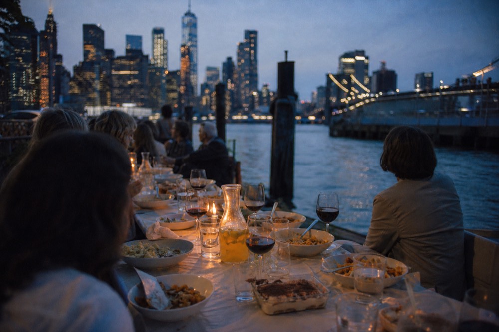 A long communal table set for a family meal with bowls of fresh pasta, a jug of juice, glasses of red wine and a dish of tiramisu in the middle