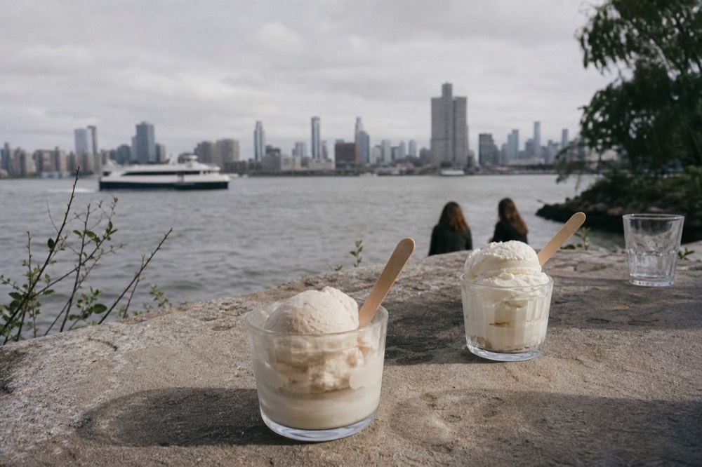 Scoops of pale gelato in small glass cups on a stone farmhouse table with a wooden spoon and afternoon shadows across the surface