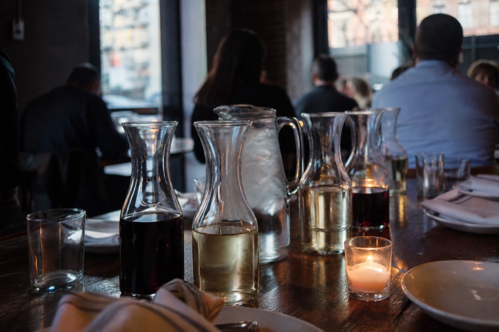 Small glass carafes of red and white wine and a jug of iced water on a long communal table set with plain white plates and linen napkins