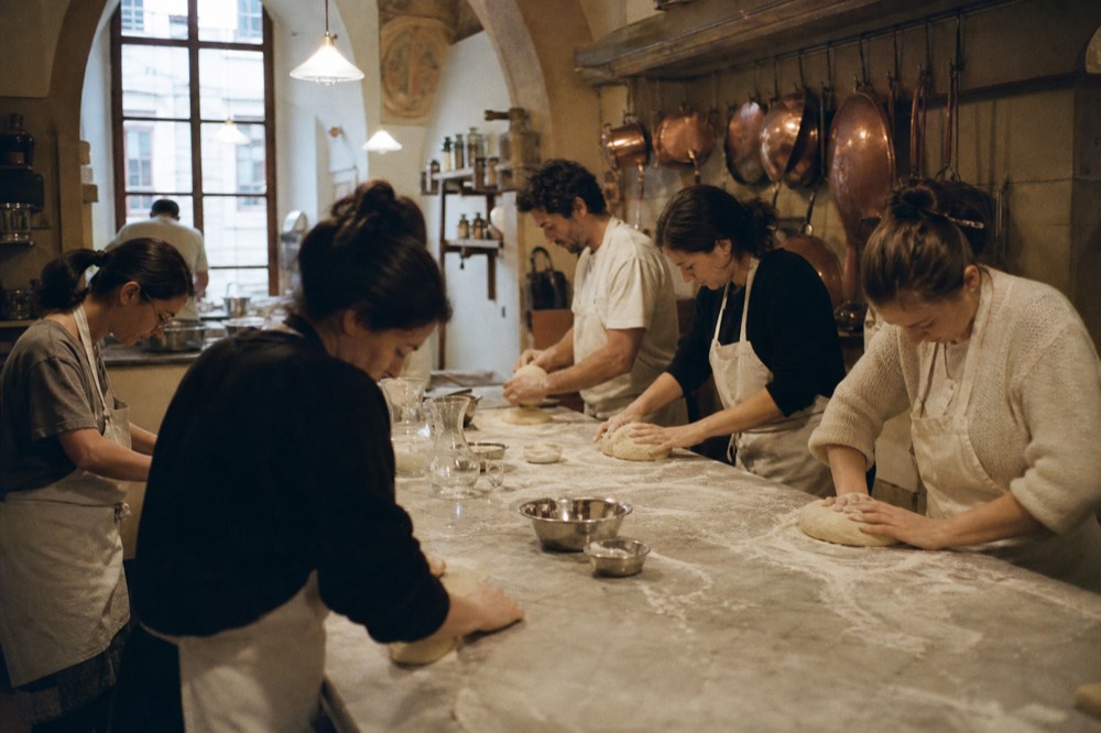Six people at a long floured bench in a vaulted Florentine kitchen, each working a separate dough, warm lamps overhead and copper pans on the wall