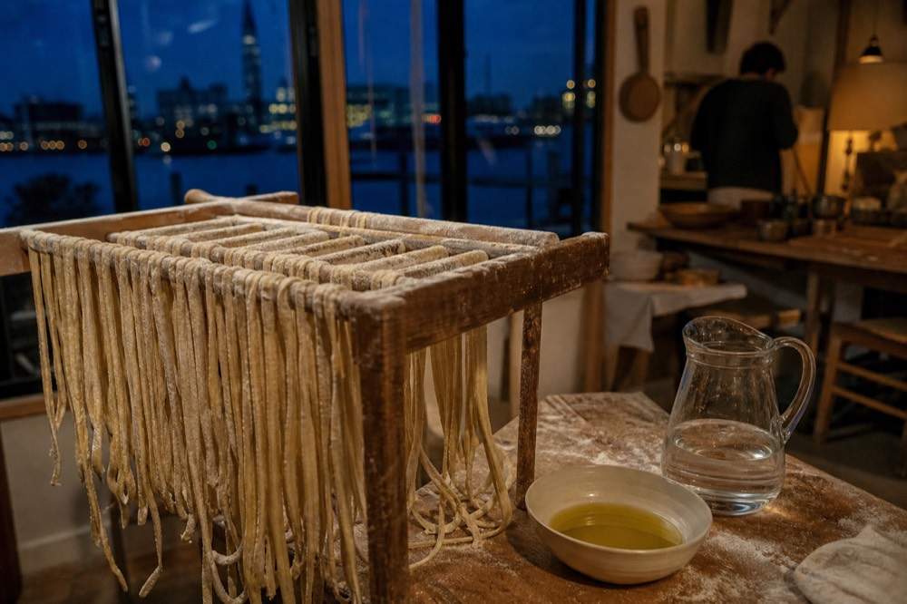 Long strands of hand-rolled pici drying on a floured wooden rack beside a small bowl of olive oil and a jug of water
