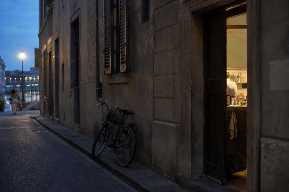 A quiet Florence side street in soft August light with shuttered windows, a parked bicycle and a single open doorway leading into a kitchen