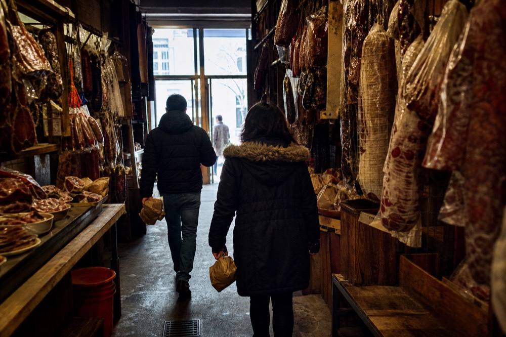 Two shoppers seen from behind carrying paper-wrapped parcels down a narrow market aisle lined with hanging cured meats