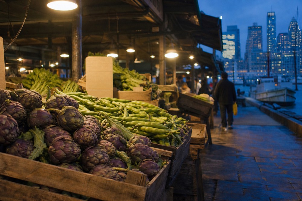 Wooden crates of purple artichokes and fava beans at a market stall, price cards blank, damp stone floor beneath