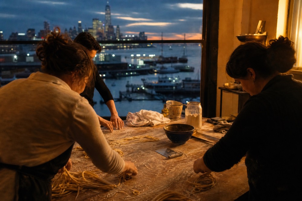 Small group of adults around a long floured table shaping thick hand-rolled pasta strands, warm kitchen light from a side window