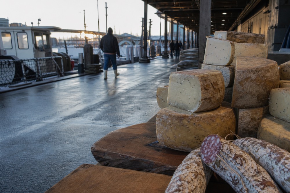 Wheels and wedges of aged sheep's-milk cheese stacked on a market counter beside fennel-flecked salami