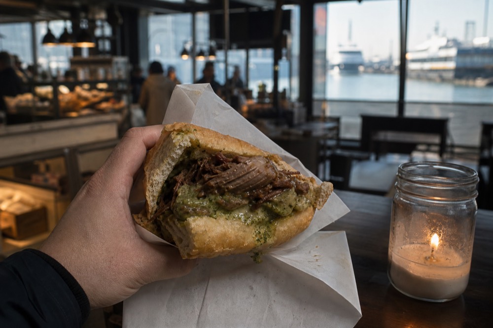Bread roll filled with slow-cooked meat and green sauce held over a paper wrapper at a market counter