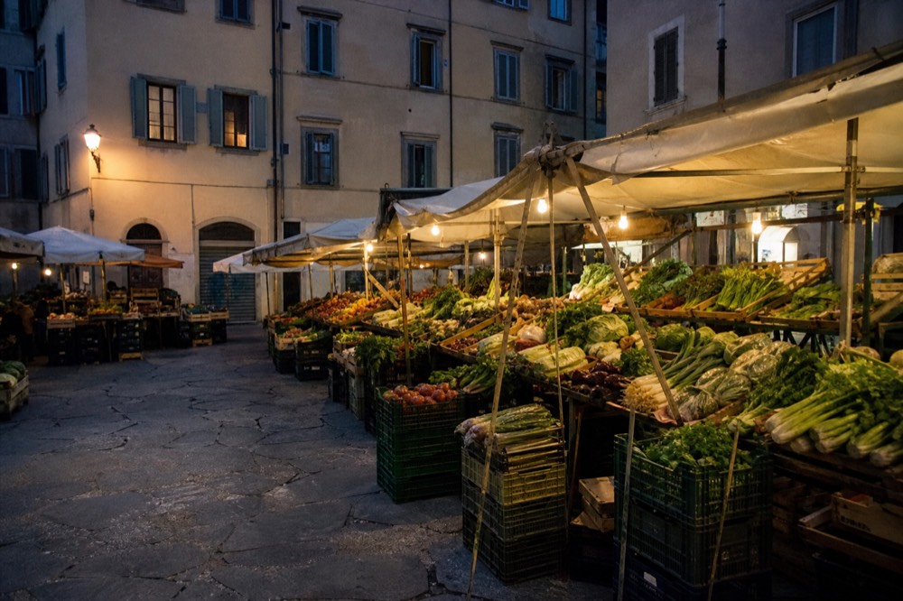 Outdoor market stalls on a quiet Florentine square, awnings over crates of vegetables, residential shutters above