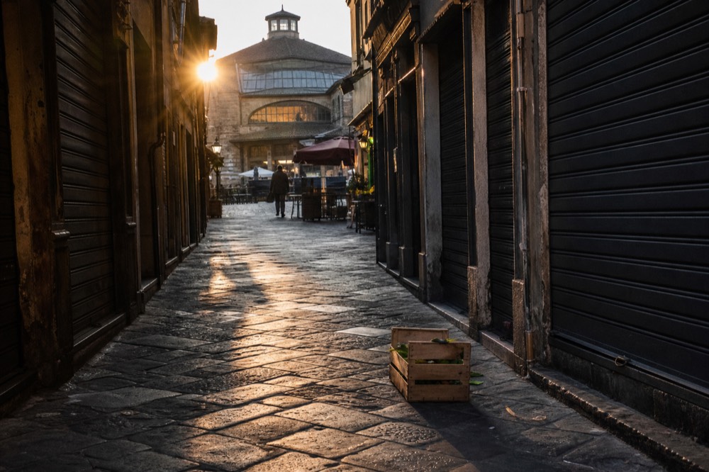 Narrow Florentine street at dawn leading toward a market building, shuttered shops and a delivery crate on the cobbles