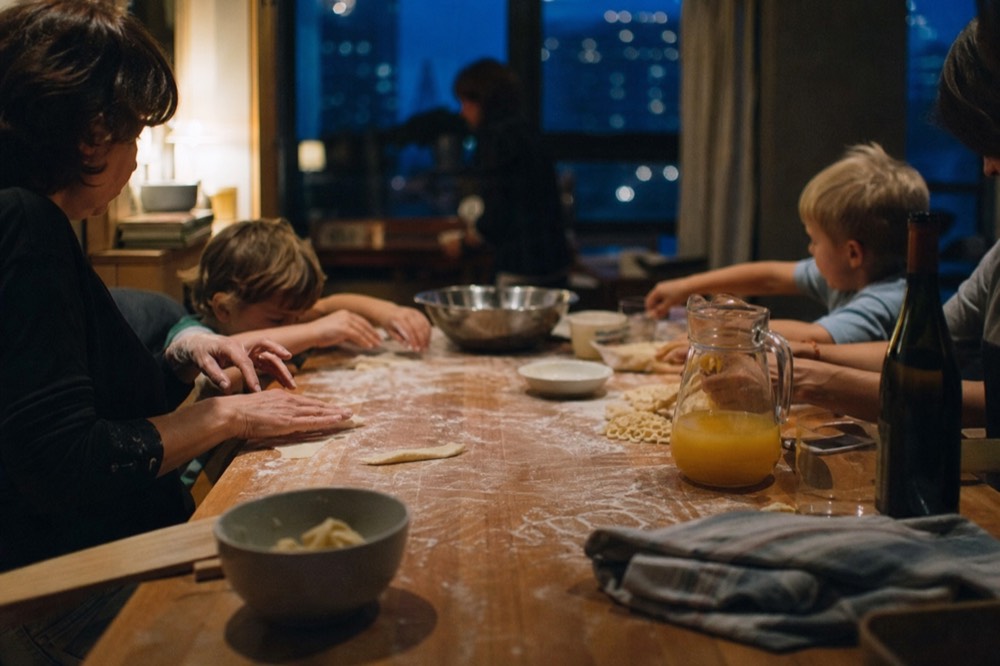 Family-sized kitchen table with small hands and adult hands shaping pasta together, a jug of juice and a bottle of wine at the end