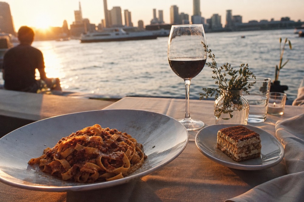 Finished plate of tagliatelle in meat sauce next to a small portion of tiramisu and a glass of red wine on a linen cloth