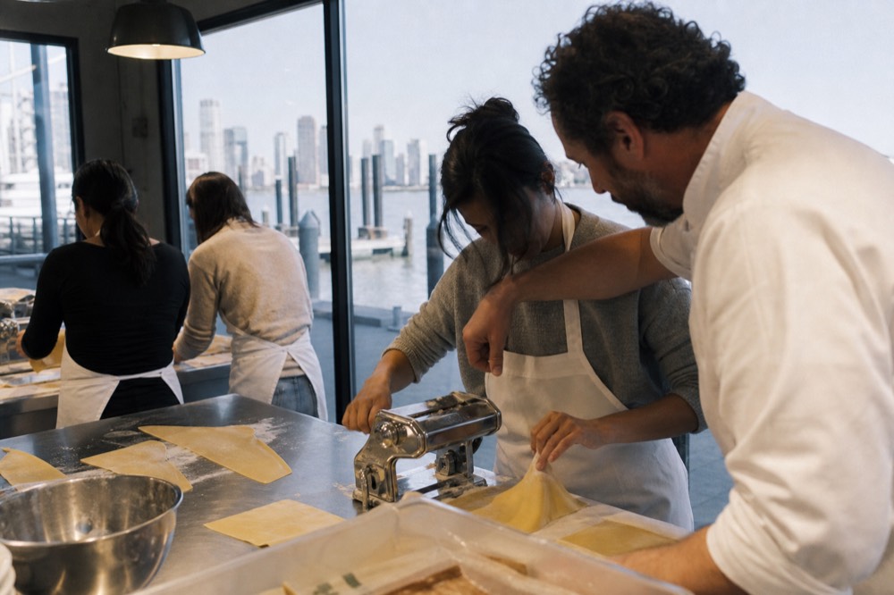 A small group of adults at a long stainless bench rolling pasta sheets, a chef leaning in to adjust one person's hands, soft overhead kitchen light