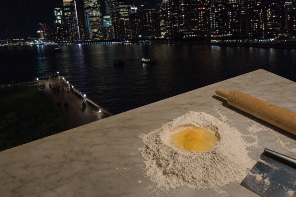 Overhead view of a marble worktop with a mound of tipo 00 flour, cracked eggs in the well, a bench scraper and a wooden rolling pin