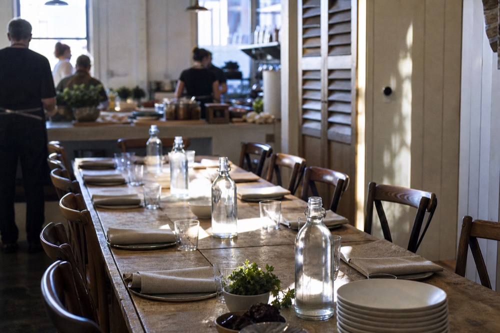 A long farmhouse table set for a private cooking lesson with linen napkins, water carafes and a stack of white plates, late afternoon light through shutters