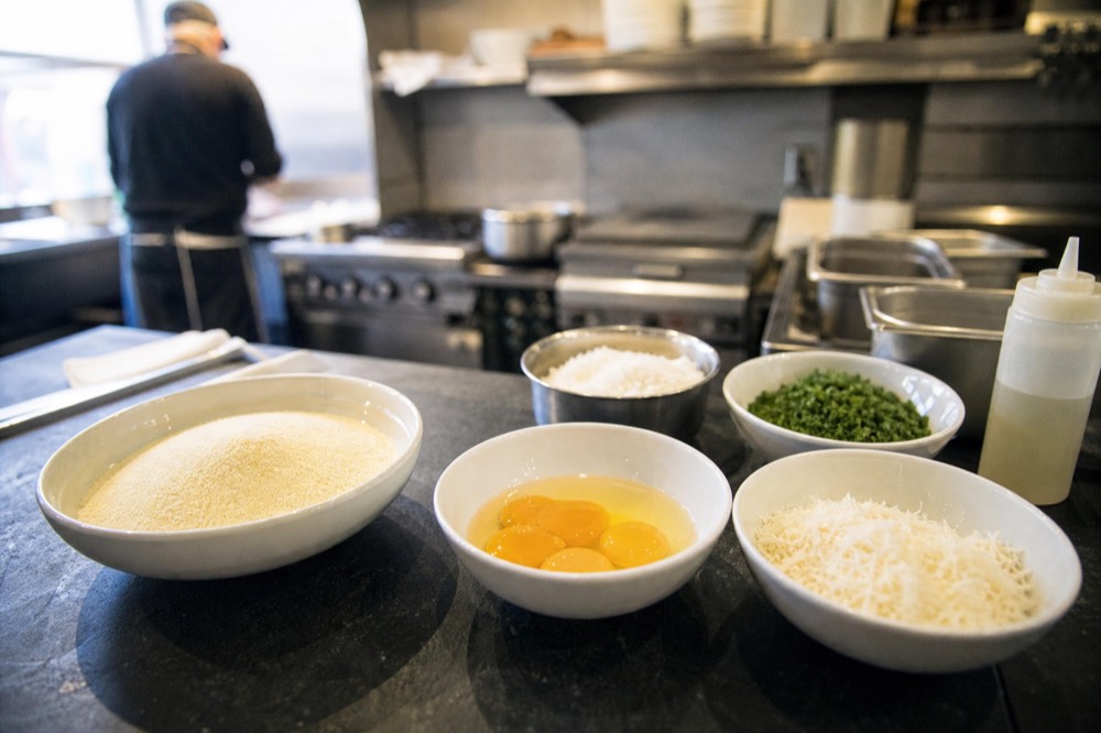 Neatly arranged mise en place bowls of semolina, egg yolks, chopped herbs and grated cheese on a dark stone counter in a small professional kitchen