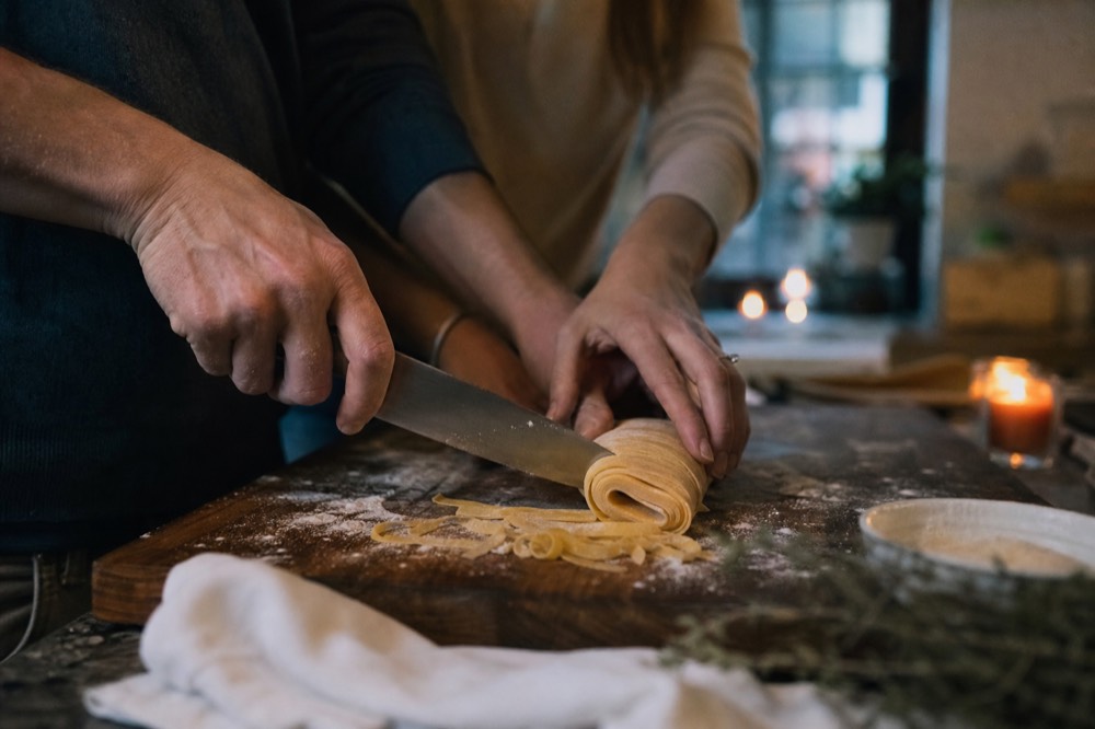 A couple's hands cutting ribbons from a rolled pasta sheet with a knife, flour dusted across a dark wood board, candle glow in the background