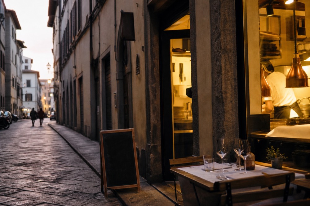 A narrow Florentine street at dusk with warm light spilling from a ground-floor kitchen window and a chalkboard easel outside