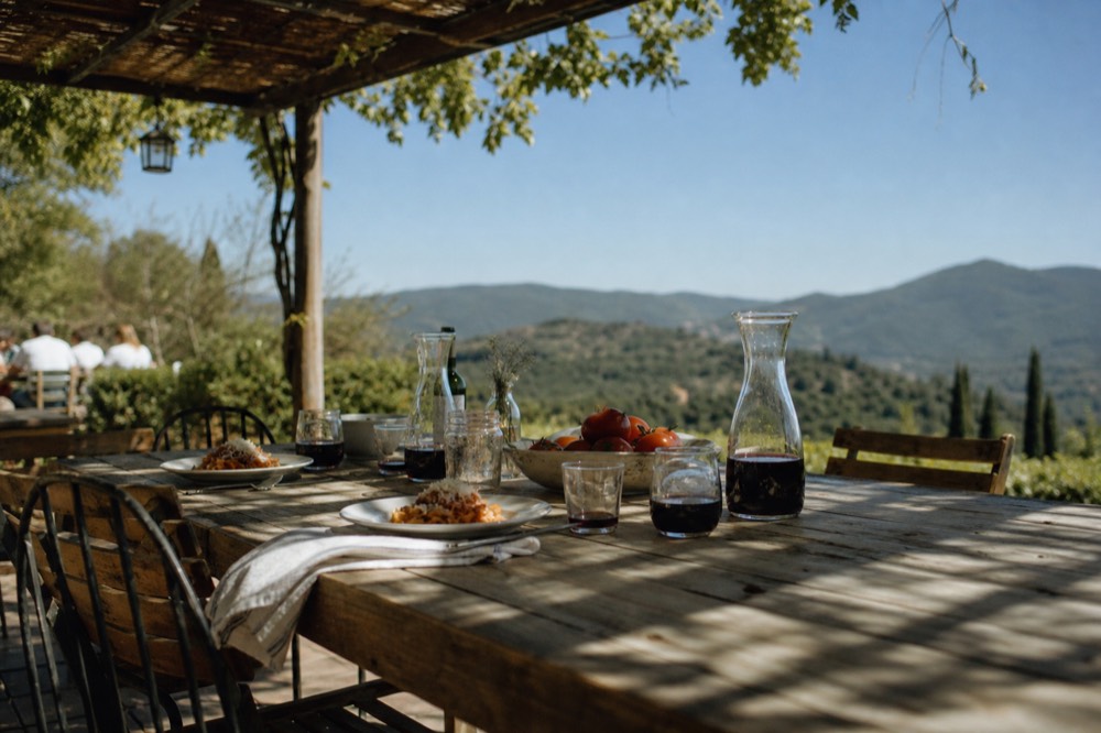 A rustic outdoor table under a pergola set with plates of pasta, carafes of red wine and a bowl of tomatoes, rolling green hills behind