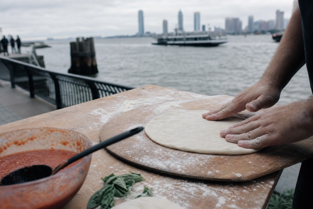 Hands pressing a round of pizza dough on a floured wooden board beside a bowl of crushed tomatoes and torn basil leaves