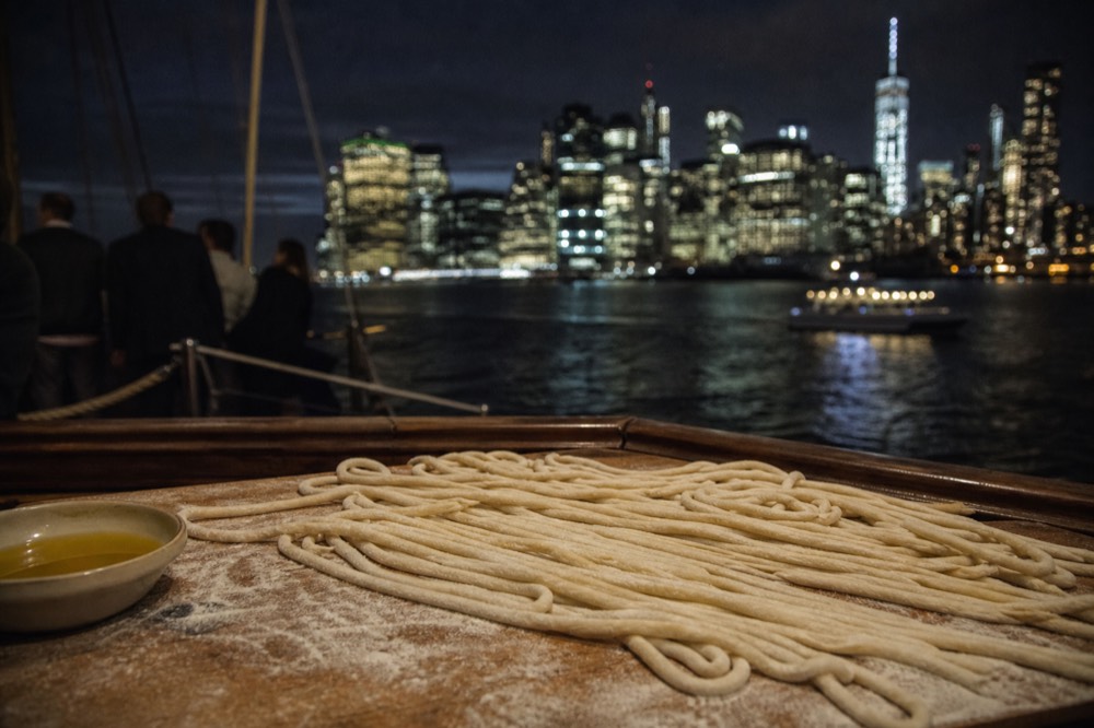 Long strands of hand-rolled pici pasta resting on a flour-dusted wooden board next to a small bowl of olive oil