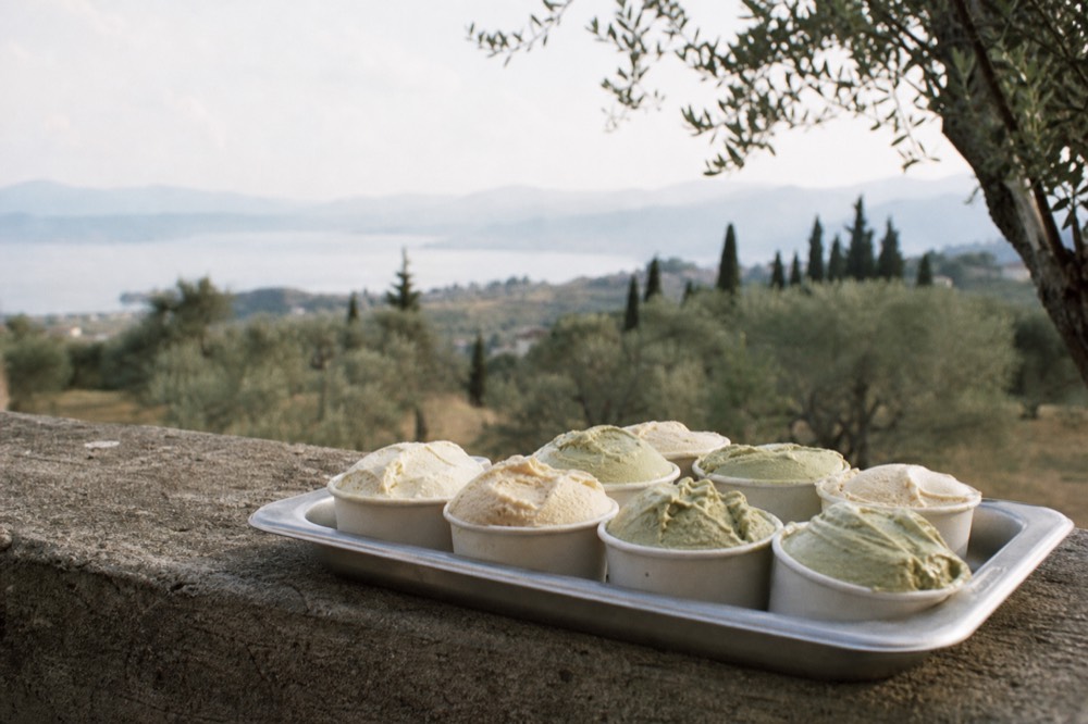 A tray of small gelato tubs in pale cream and pistachio shades on a stone ledge, an olive grove blurred in the background